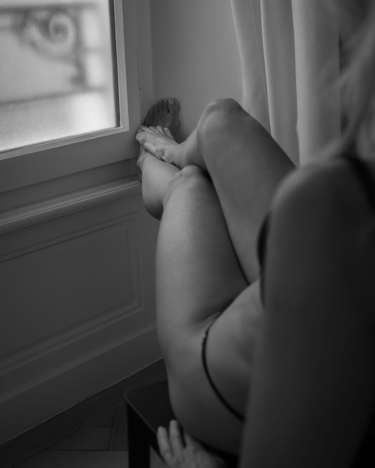 portraits intimistes noir et blanc photographié par une femme. Femme assise sur une chaise ayant les pieds posés sur le rebord intérieur de la fenêtre en bois fermée. On voit la femme de dos et principalement ses jambes et uniquement une petite partie de l'arrière de son bras gauche, une partie de son ventre ainsi que quelques doigts de sa main gauche.