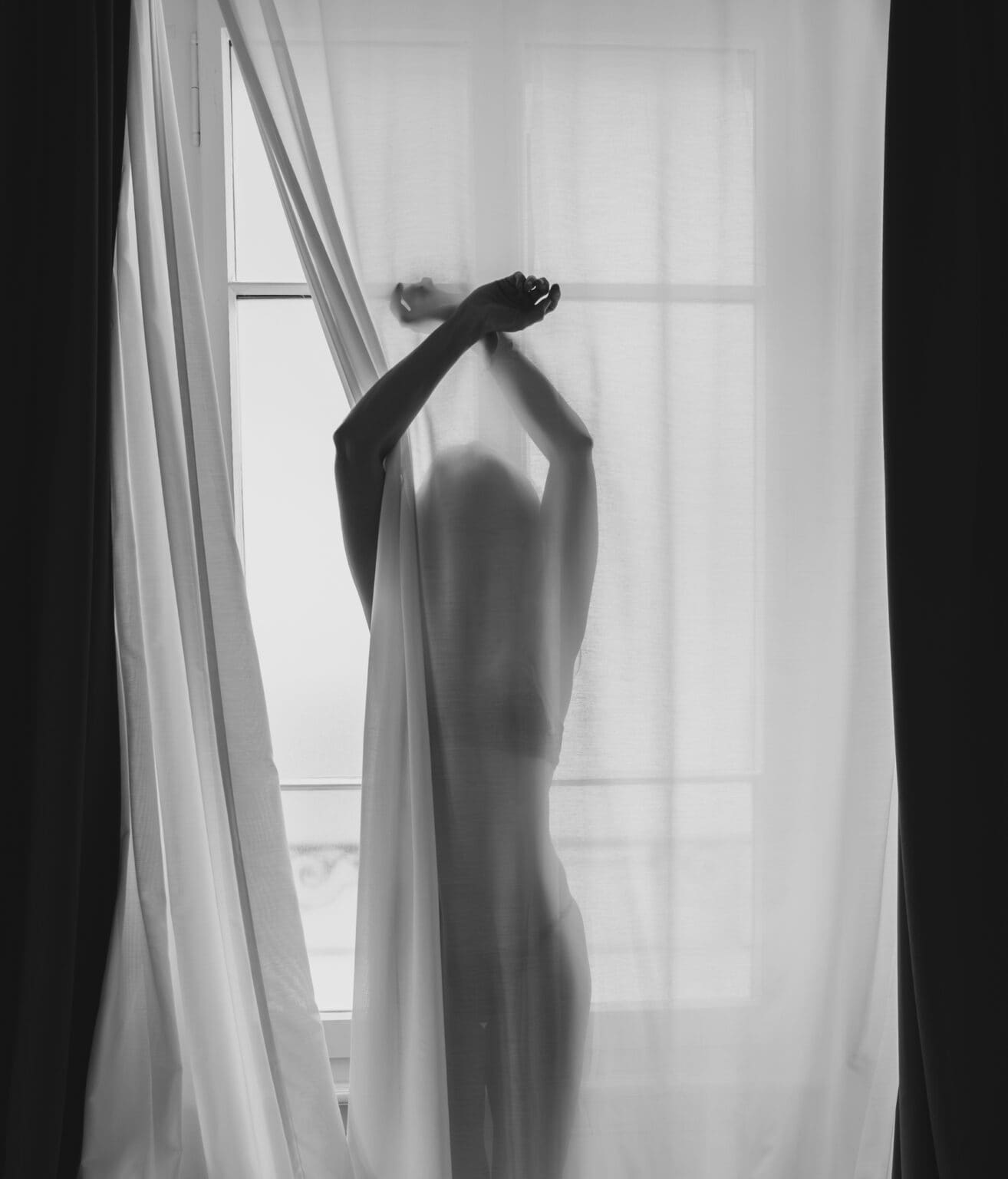 Portrait artistique intimiste en noir et blanc d'une femme en sous-vêtements noirs, se tenant derrière un rideau en transparence. Elle est est à contre-jour. Elle passe son bras droit devant le rideau
