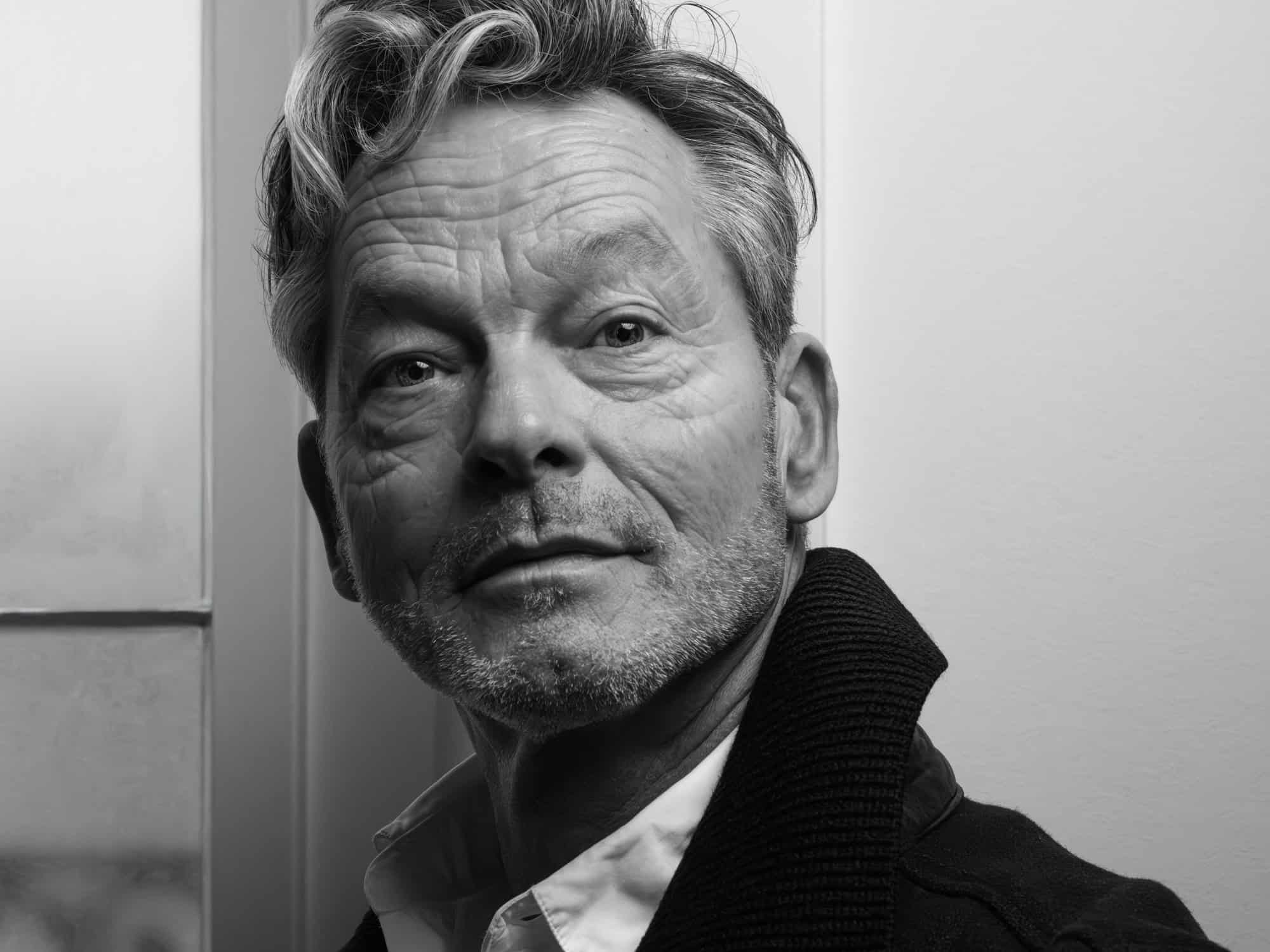 Portrait professionnel noir et blanc. Gros plan sur son visage et regard d'un homme de plus de 60 ans, ridé, cheveux poivre et sel. Il regarde de manière frontale l'objectif.