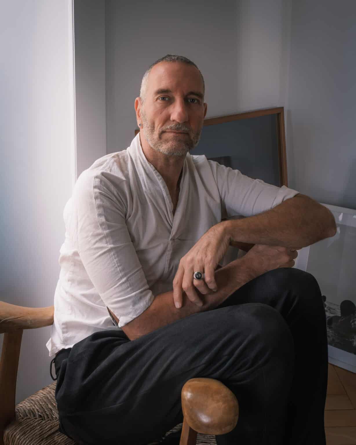 Portrait professionnel couleur. Portrait photographié par une femme. Il s'agit d'un homme assis de côté sur une chaise avec des accoudoirs. L'homme porte une chemise blanche et un pantalon noir. Il porte chevalière sur la main gauche. Pn devine des cadres appuyés contre le mur à l'arrière.