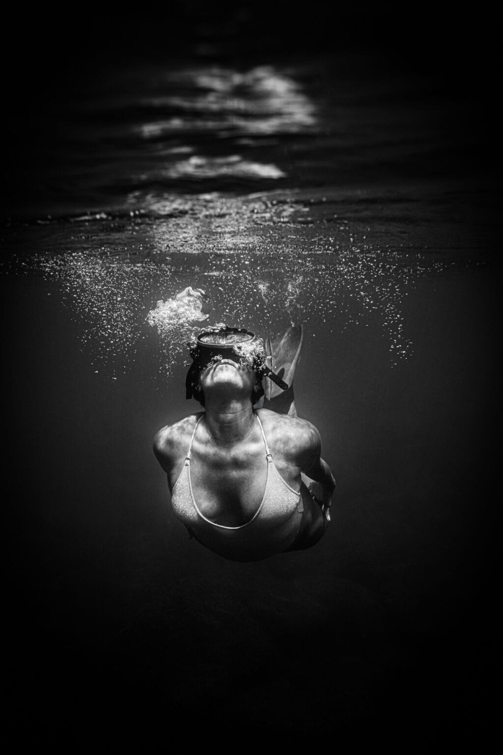 Portrait underwater en noir et blanc. Femme juste en-dessous de la surface. Corps en U, les bras le long du corps. Visage et masque regardant la surface. Femme expire, beaucoup de bulles.