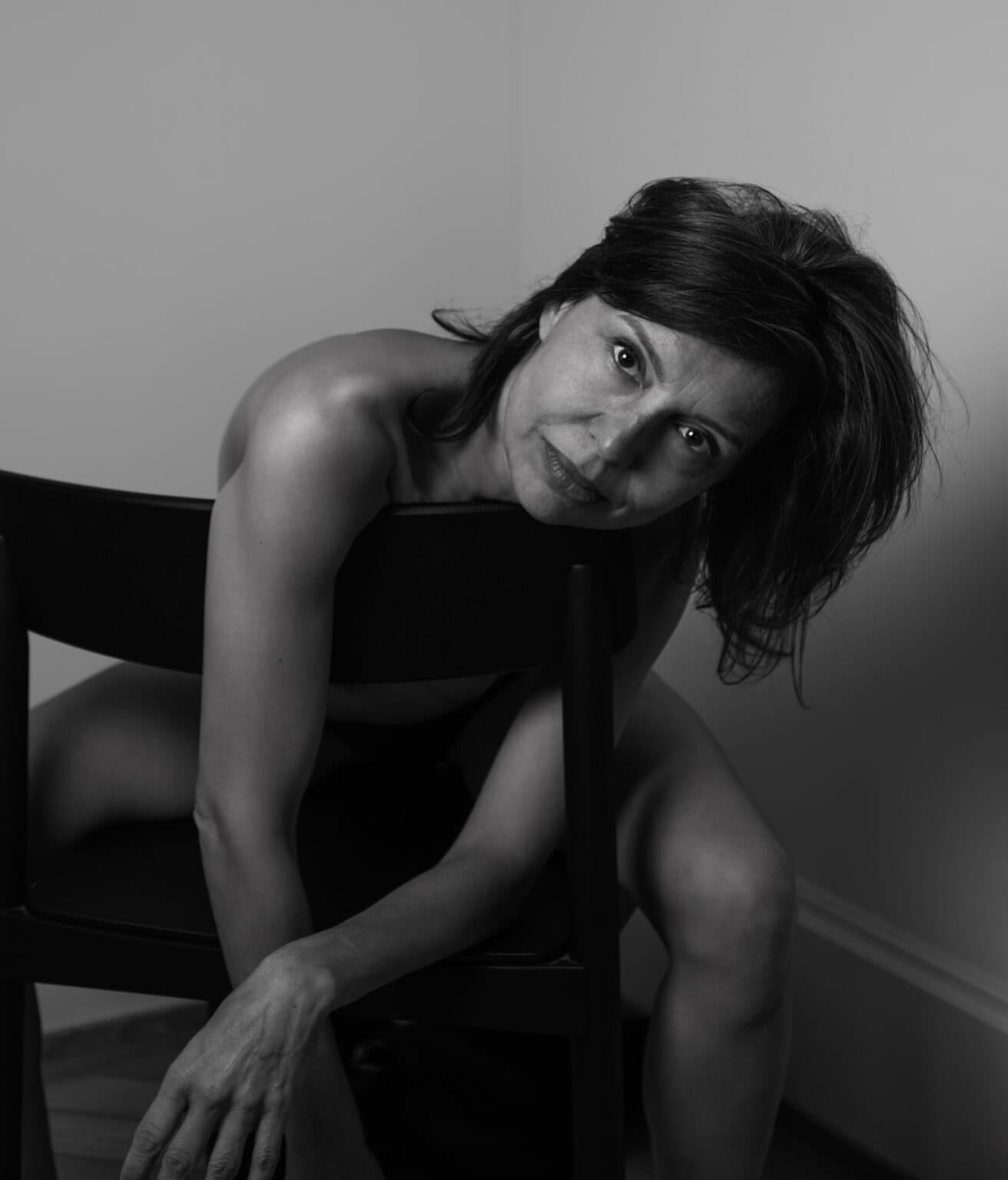 nu artistique feminin en noir et blanc, portrait intimiste photographié par une femme. Femme nue assise sur une chaise. On devine nudité, mais on voit les jambes, épaules dénudés uniquement. Elle regarde avec intensité.