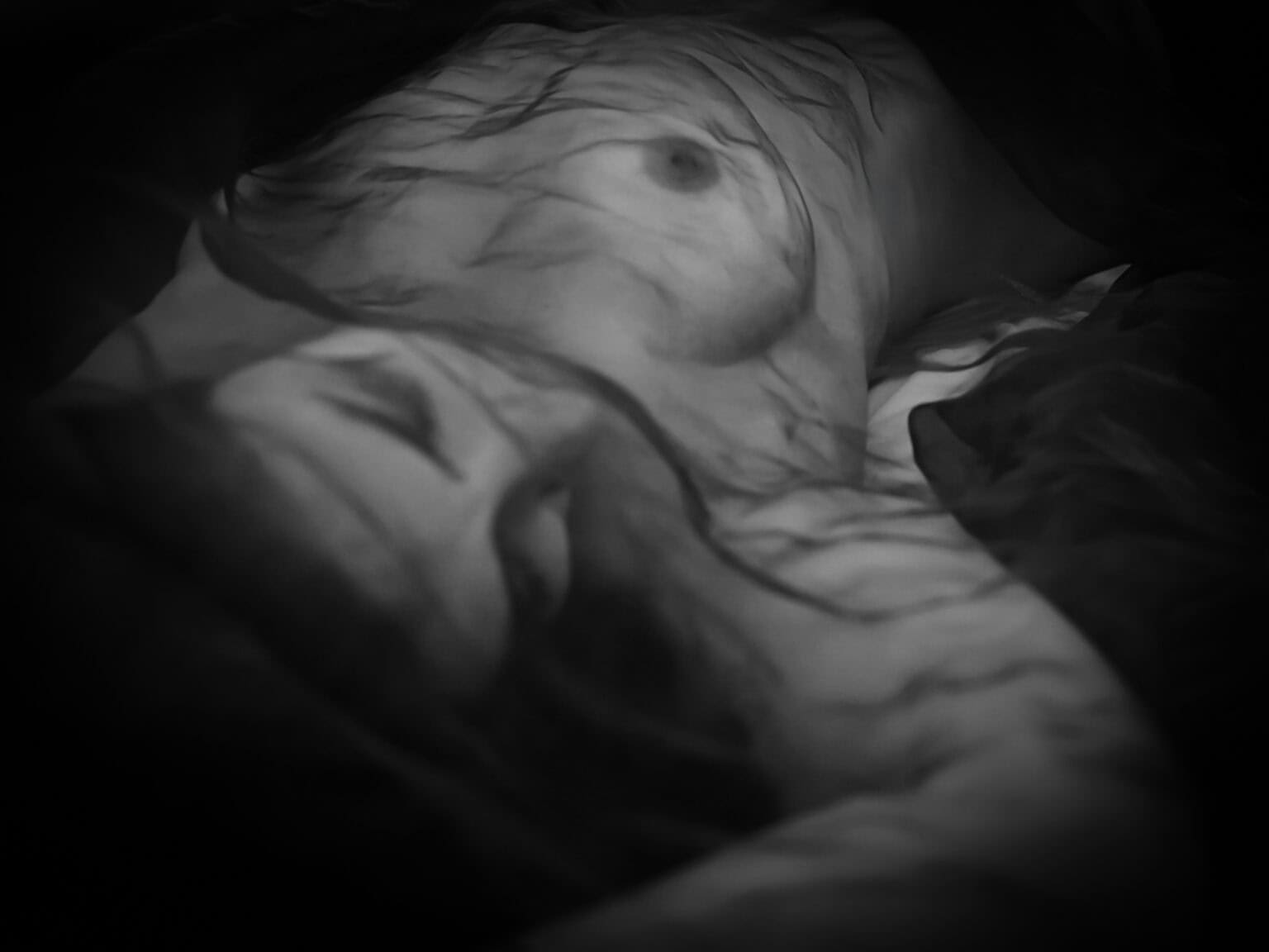 nu artistique feminin en noir et blanc – portrait intimiste photographié par une femme. Femme allongée sur le dos. Elle porte une mousseline sur le corps et visage, donc on devine sein et visage, mais cela reste flou