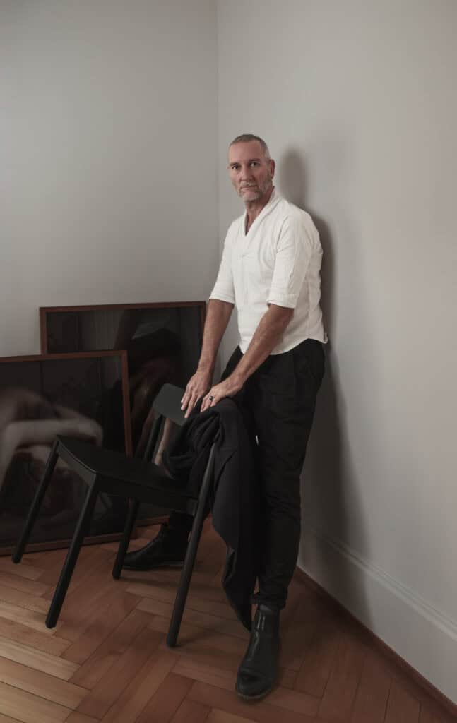 Portrait professionnel couleur. Homme se tenant apuyé contre mur avec mains posées sur chaise noire. Il porte un pantalon noir et une chemise blanhce,