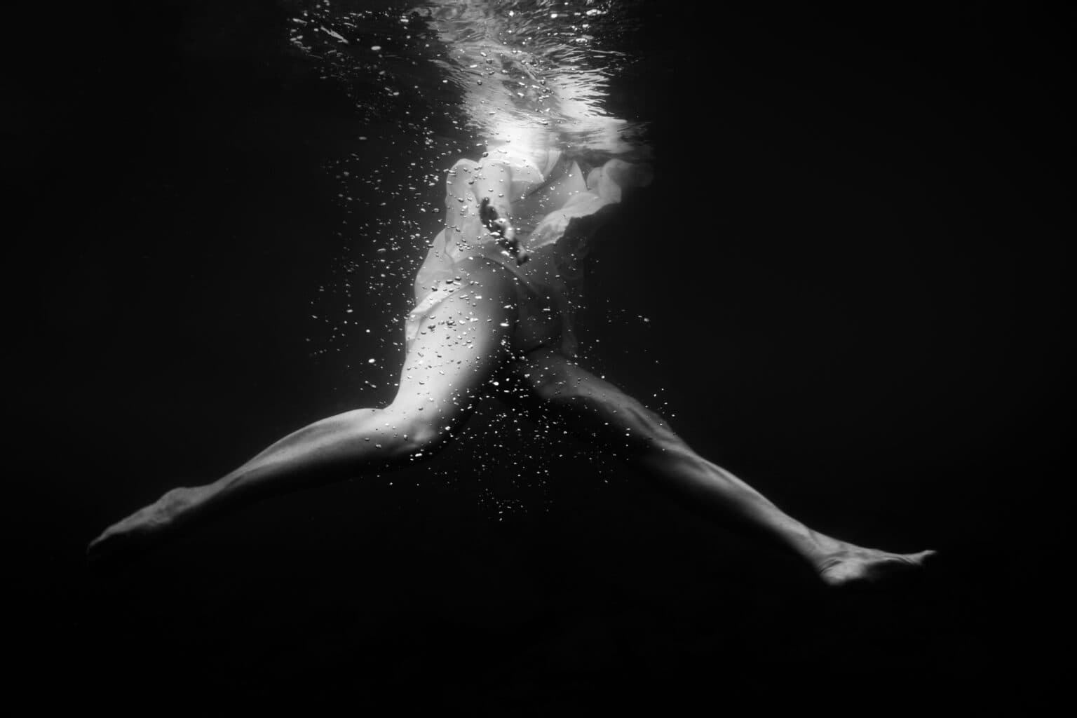Portrait underwater noir et blanc. La photo a été prise de profil, prise un peu depuis le bas. Il s'agit d'une femme portant une chamise blanche écartant les jambes dans le sens longitudinal. Elle a tout le coprs immergé sauf la tête.