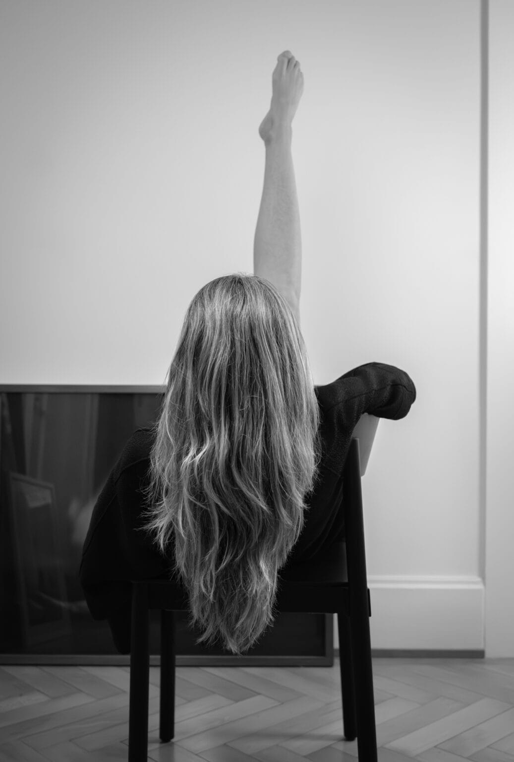 Portrait artistique créatif en noir et blanc. Photographié par une femme. Femme assise de dos sur une chaise, longue chevelure blonde. Elle lève un seule jambe.