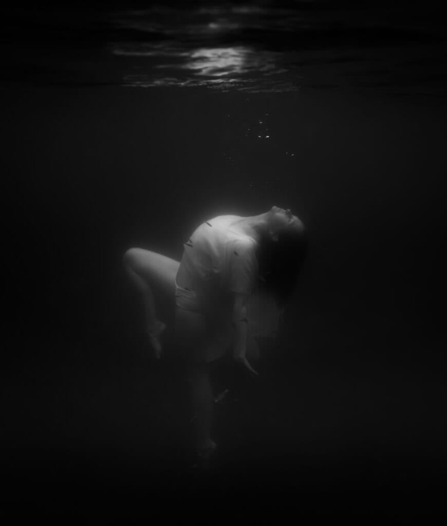 Portrait underwater noir et blanc. portrait photographié par une femme. Il s'agit d'une femme prise de profil qui remonte à la surface. Elle est habillée d'une robe blanche. La photo est sombre, seul le haut du corps, visage et jambe droite repliée sont dans la lumière