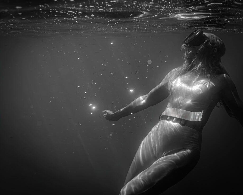Portrait underwater en noir et blanc. Femme qui est juste sous la surface, qui regarde la surface avec un masque vintage. Elle porte un maillot gris argenté avec une ceinture de plomb. Le corps est tourné vers l'objectif, mais la femme regarde la surface. Nombreuses particules flottantes, bcp de petites tahces de lumière.