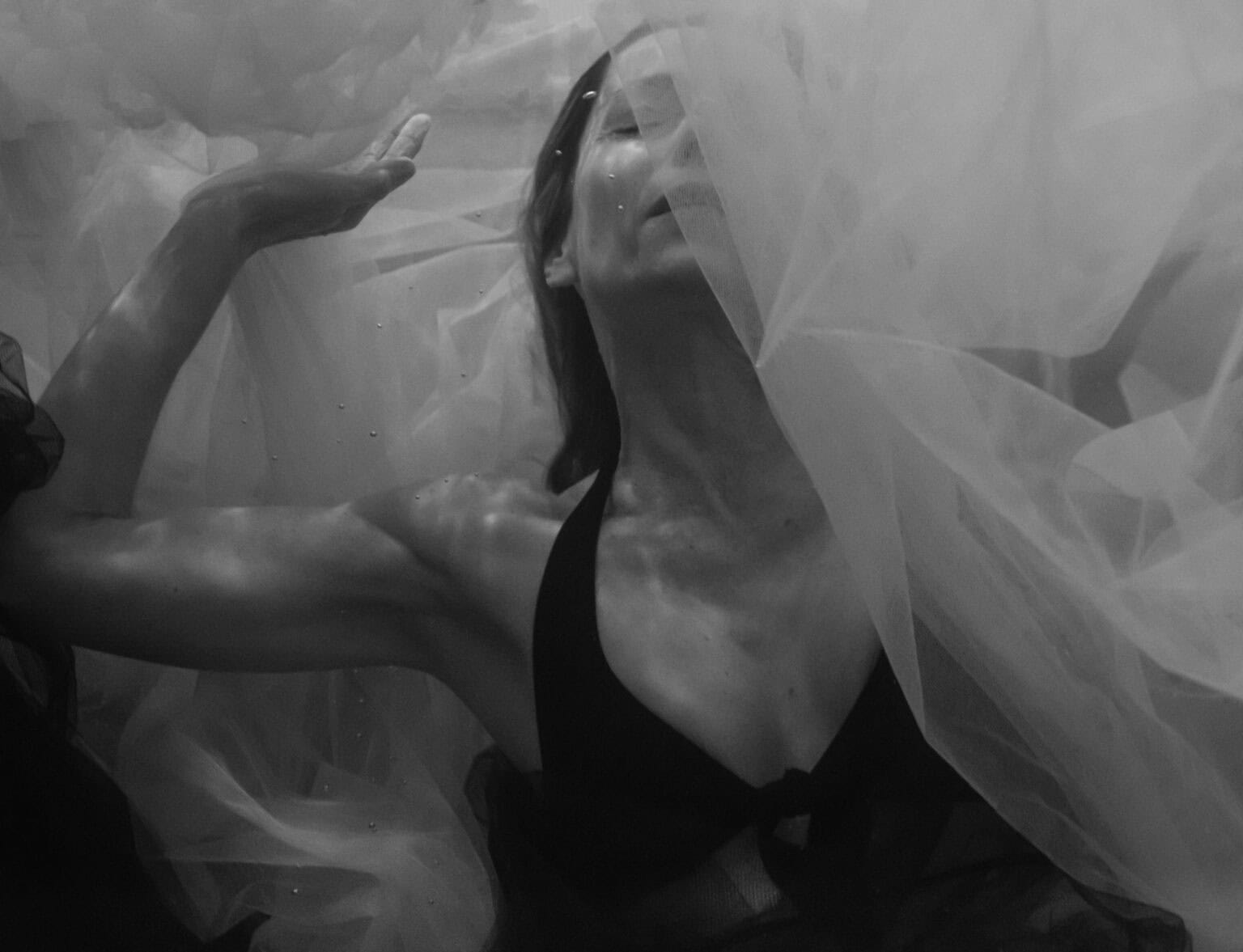 Portrait underwaternoir et blanc. portrait photographié par une femme. Femme qui se trouve derrière et en dessous de mousseline blanche. ELle a bikini noir. On ne voit que le côté droite du corps, avec le bras levé droit.