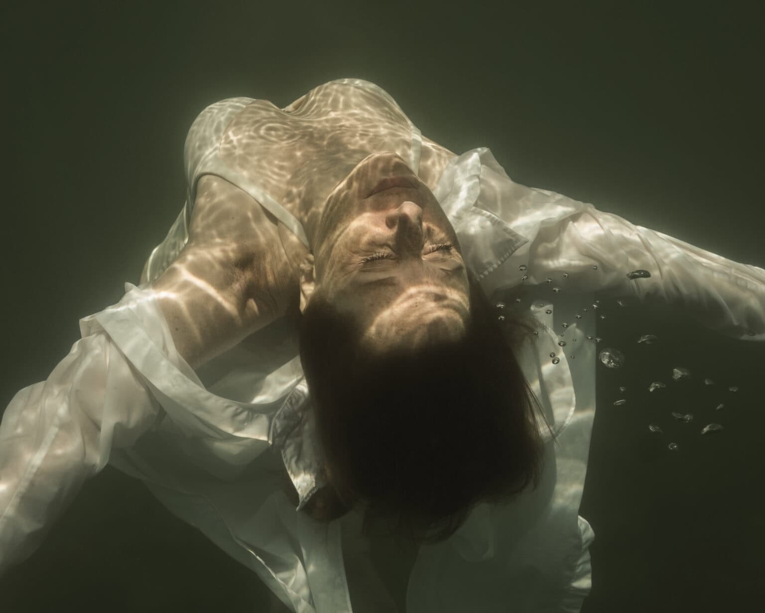 Portrait underwateren couleur. Femme en bikini blanc avec une chemise déboutonnée. La photo est prise de derrière et haut la femme qui remonte à la surface, les bras ouverts. On voit le visage avec les yeux fermés ainsi que les début de ses bras et le heut de son buste.
